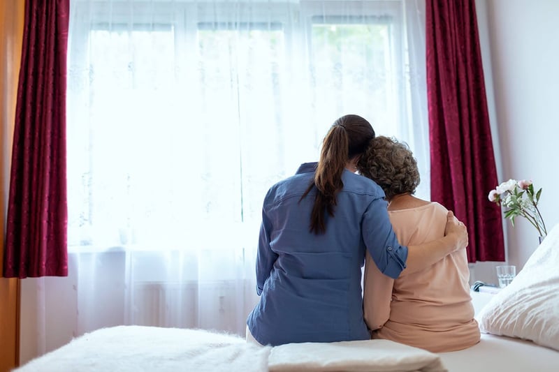 Older woman with younger woman sitting on bed staring out the window